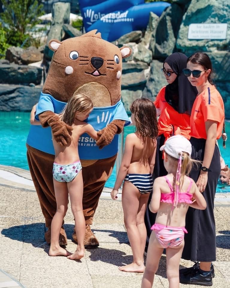 Ein Biber-Maskottchen in einem blauen T-Shirt steht neben einer Gruppe von Kindern in Badeanzügen in einem Wasserpark.