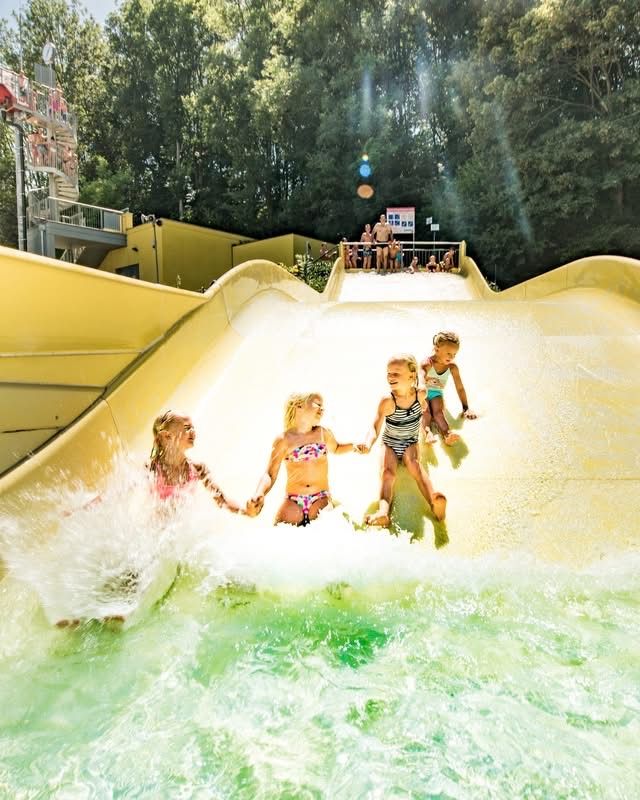 Vier Mädchen rutschen an Händen haltend eine gelbe Wasserrutsche hinunter. Dahinter beobachten Leute von einer Plattform aus. Bäume und ein Gebäude sind im Hintergrund.