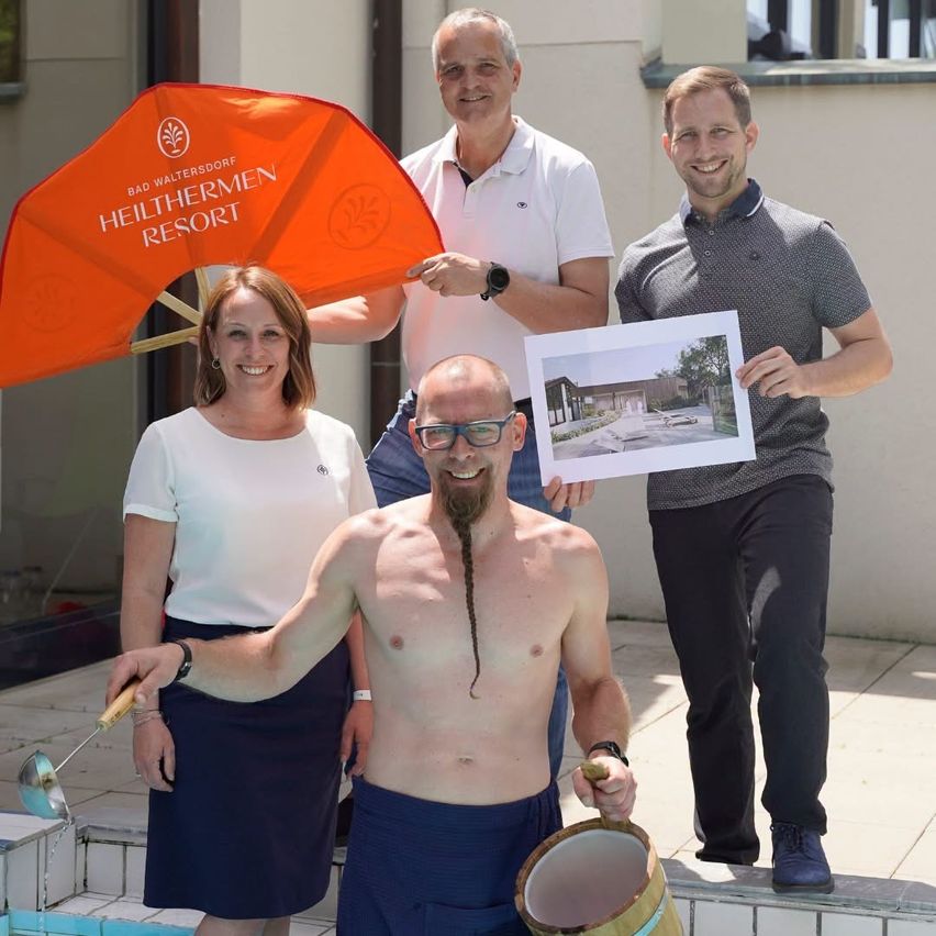 Drei Personen stehen am Pool, eine hält ein oranges Banner für Bad Waltersdorf Heuthermen Resort, eine andere hält eine Zeichnung und die dritte einen Becher.