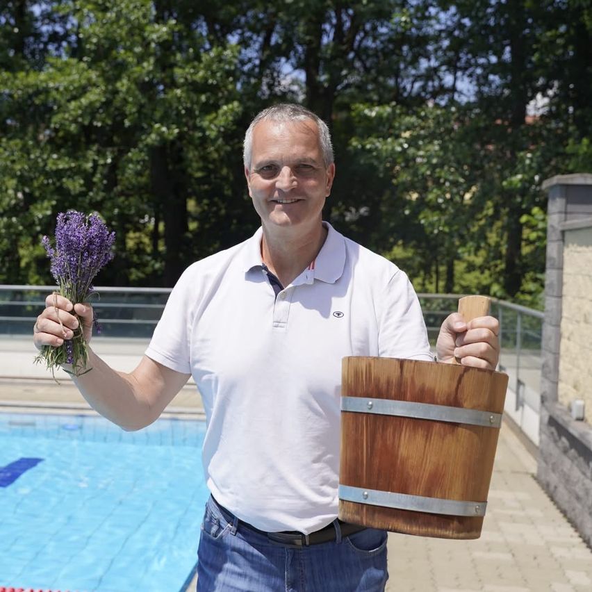 Ein Mann hält einen Lavendelstrauß und einen Holzeimer in der Hand und steht lächelnd am Pool.