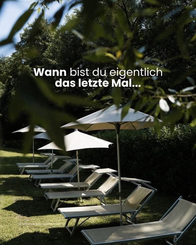 Eine Reihe von Liegestühlen auf einer Wiese mit weißen Sonnenschirmen. Ein deutscher Satz, 'Wann bist du eigentlich das letzte Mal...', ist auf dem Bild geschrieben.