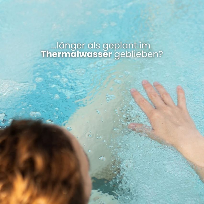 Eine Person ist in einem Pool, teilweise eingetaucht. Die Bildunterschrift lautet: '...länger als geplant im Thermalwasser geblieben?' Das Wasser ist klar.
