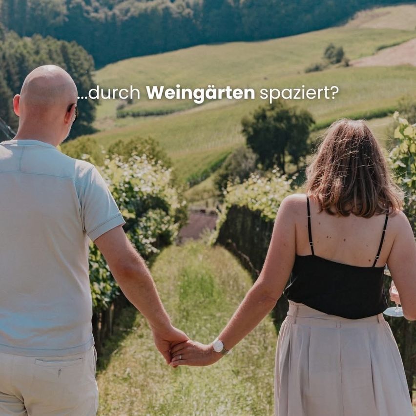 Ein Paar geht Hand in Hand durch einen Weinberg mit Blick auf ein grünes Feld und Hügel in der Ferne.