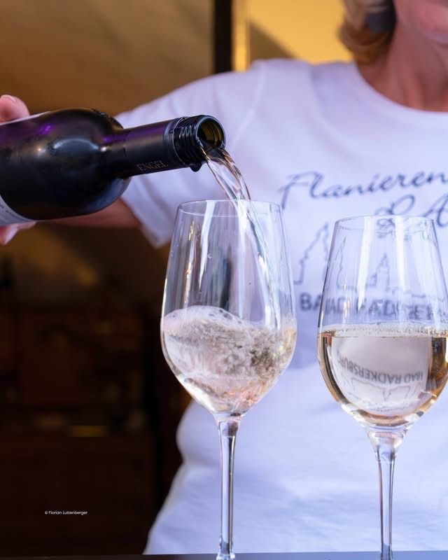 Eine Frau gießt Wein aus einer Flasche in zwei Gläser. Der Wein ist prickelnd und sieht schaumig aus. Der Hintergrund ist verschwommen.