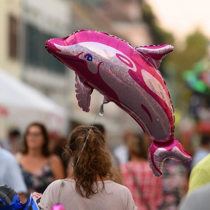 Eine Frau trägt einen rosa Delfinballon. Im Hintergrund sind mehrere Menschen in einer verschwommenen Straßenumgebung.