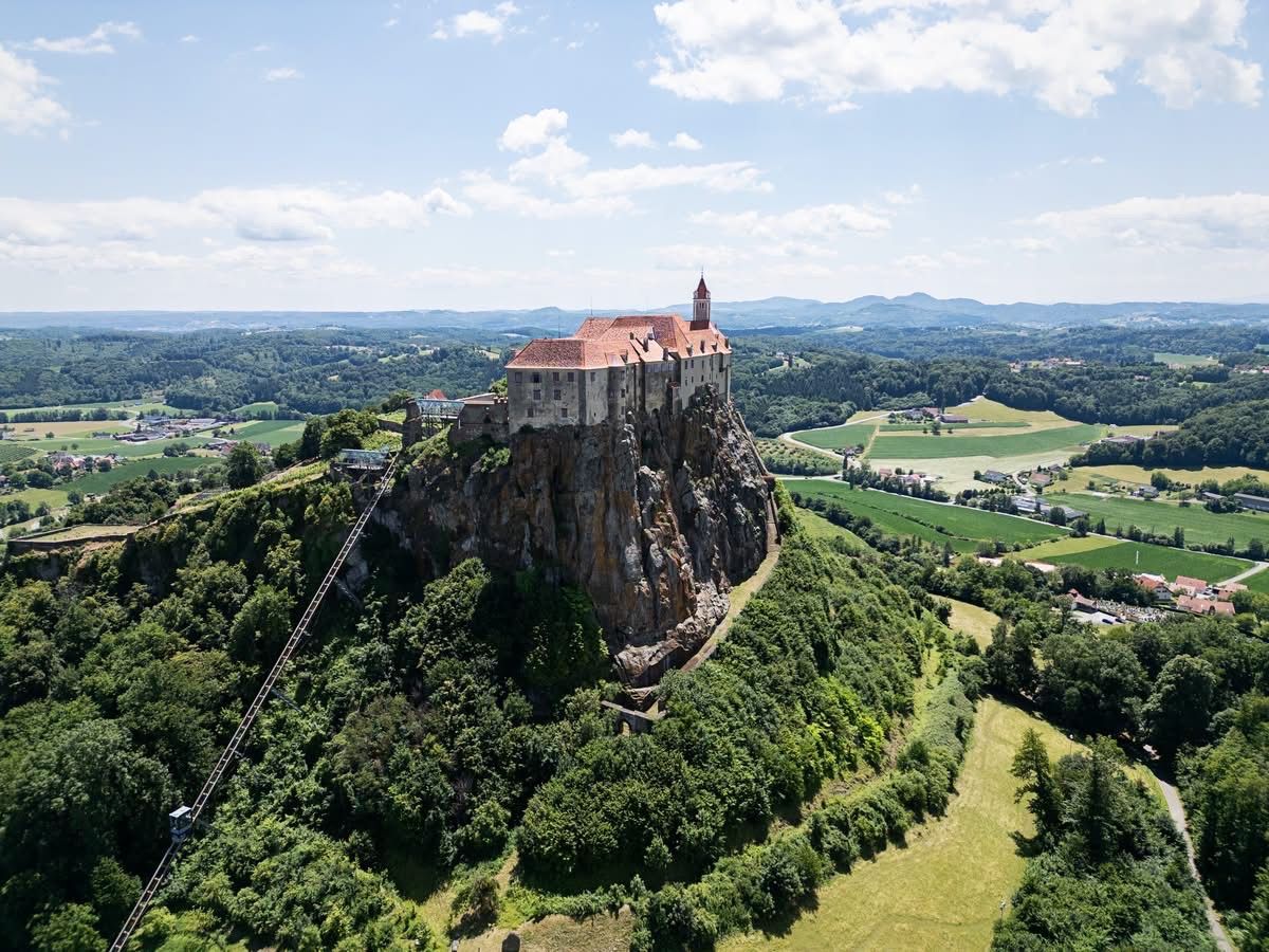 An aerial view of a medieval castle perched atop a rocky cliff, surrounded by lush greenery and a distant mountain range under a partly cloudy sky.