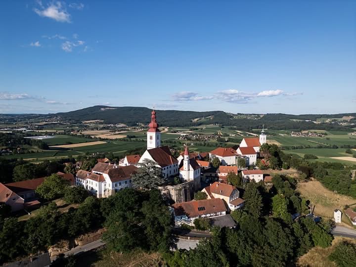 An aerial view of a village with a church at its center, surrounded by lush green fields and trees, under a clear blue sky.