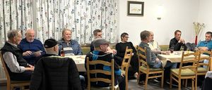 Eine Gruppe von Menschen sitzt um einen Tisch in einem Raum mit einem Blumenvorhang, einem Bilderrahmen und einer beleuchteten Lampe. Einige haben Getränke, andere schauen etwas an.