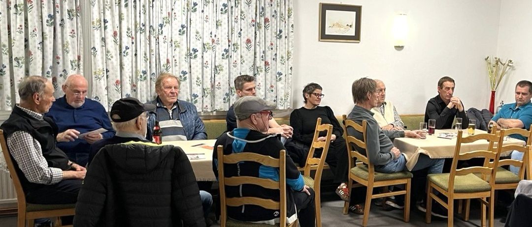 Eine Gruppe von Menschen sitzt um einen Tisch in einem Raum mit einem Blumenvorhang, einem Bilderrahmen und einer beleuchteten Lampe. Einige haben Getränke, andere schauen etwas an.