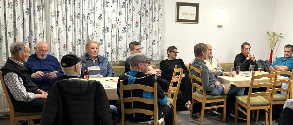 Eine Gruppe von Menschen sitzt um einen Tisch in einem Raum mit einem Blumenvorhang, einem Bilderrahmen und einer beleuchteten Lampe. Einige haben Getränke, andere schauen etwas an.