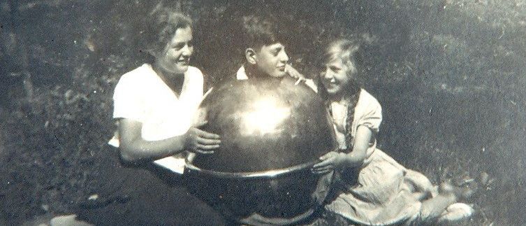Drei Kinder, zwei Mädchen und ein Junge, sitzen auf Gras mit einer großen Metallkugel zwischen ihnen und lächeln.