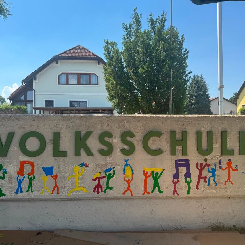 Eine Betonmauer mit einem farbenfrohen Wandbild von Menschen, die Gegenstände wie Bücher und Stifte halten. Das Wort 'VOLKSSCHUL' ist darauf geschrieben. Dahinter steht ein weißes Haus mit Bäumen.