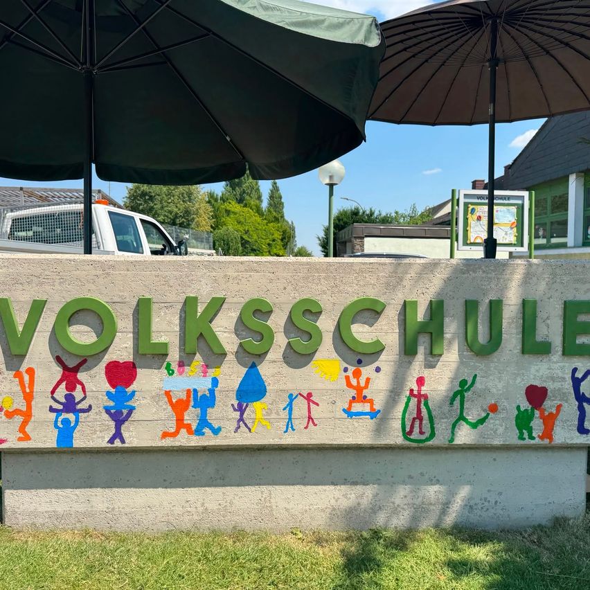 Eine Betonwand zeigt das Wort 'VOLKSSCHULE' mit farbenfrohen Strichmännchen-Illustrationen von Kindern und Objekten. Zwei Regenschirme sind über der Wand, und ein Pick-up-Truck ist in der Ferne geparkt.