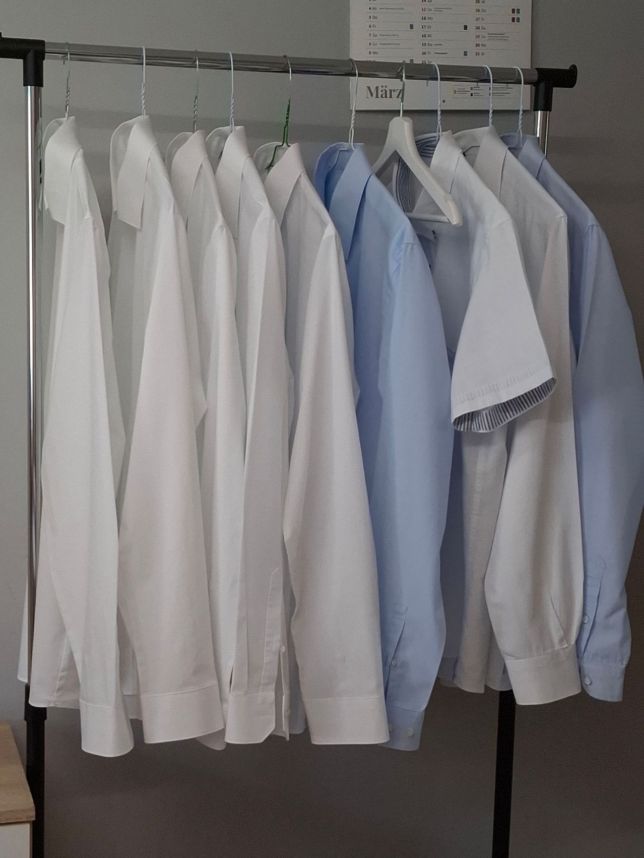 Ein Kleiderständer mit Hemden ist auf einem Metallständer ausgestellt. Die Hemden sind größtenteils weiß mit einigen hellblauen, alle ordentlich gefaltet und an weißen Kleiderbügeln hängend.