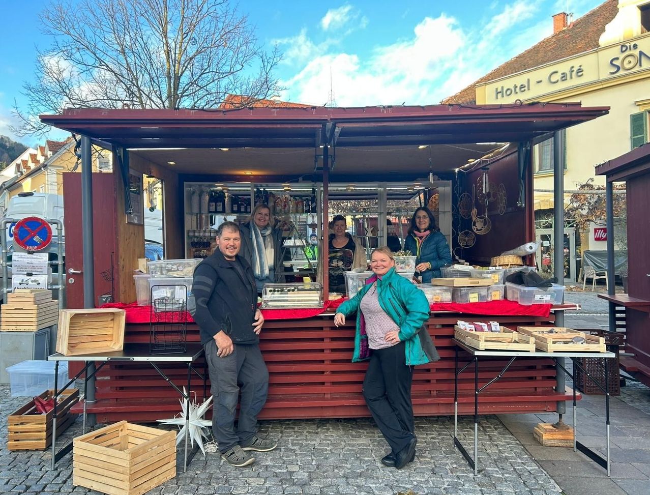 Eine Gruppe von Menschen, zwei Männer und zwei Frauen, steht vor einem roten Imbisswagen. Dahinter befinden sich mehr Menschen im Wagen, die Gegenstände vorbereiten. Holzkisten und Tische sind draußen aufgestellt.