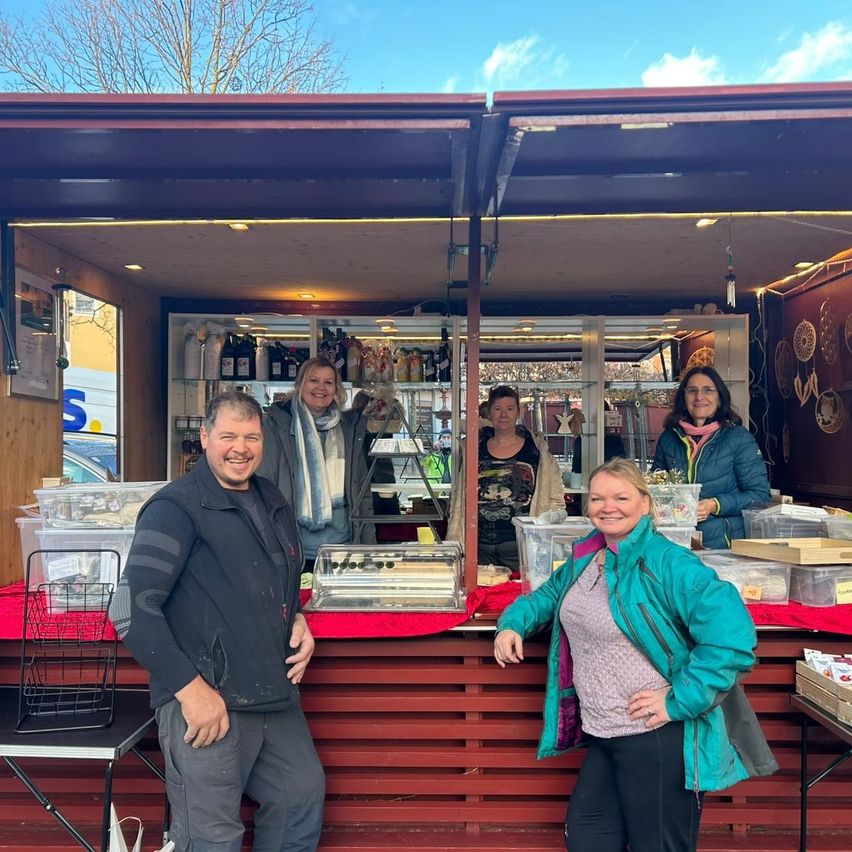 Fünf Personen stehen vor einem Essensstand an einem sonnigen Tag. Hinter ihnen sind Regale mit verschiedenen Produkten. Der Stand hat einen roten Tresen und ist mit Lichtern dekoriert.