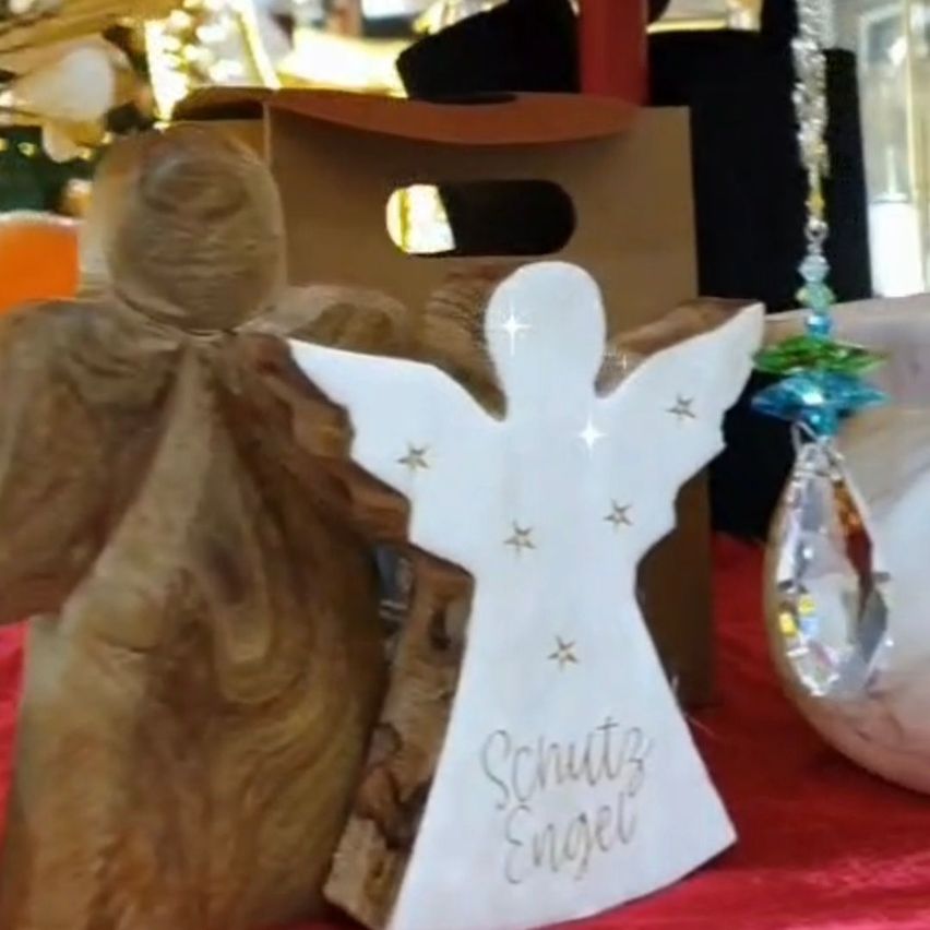 Ein hölzerner Engelschmuck mit weißem Körper, blauen Sternen und dem Aufdruck 'Schutz Engel' auf rotem Tuch.