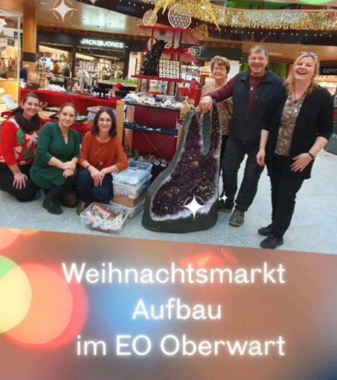 Fünf Personen posieren für ein Foto auf einem Weihnachtsmarkt mit einem großen Amethyst-Kristall. Sie lächeln und stehen oder knien um den Kristall herum. Ein Schild im Vordergrund lautet 'Weihnachtsmarkt Aufbuu im EO Oberwart.'