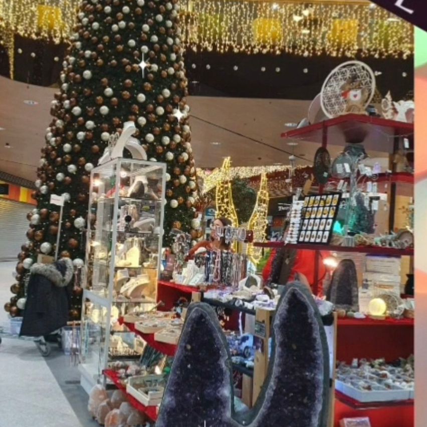 Innenansicht eines Einkaufszentrums mit einem großen Weihnachtsbaum, verziert mit Kugeln und Lichtern, vor dem ein Stand mit verschiedenen Artikeln wie Kristallen, Schmuck und Ornamenten steht.