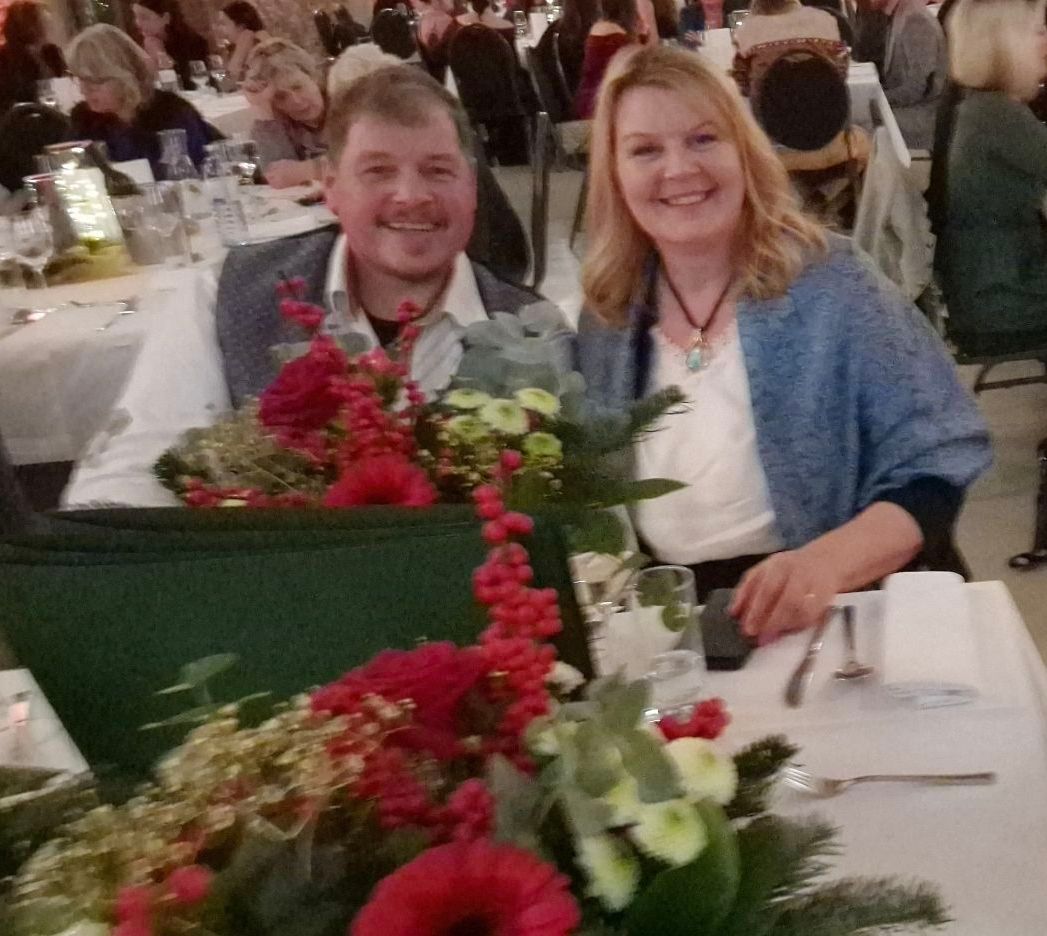 Ein Mann und eine Frau sitzen lächelnd an einem Tisch mit Blumen. Sie sind von anderen Leuten umgeben, die an Tischen sitzen.