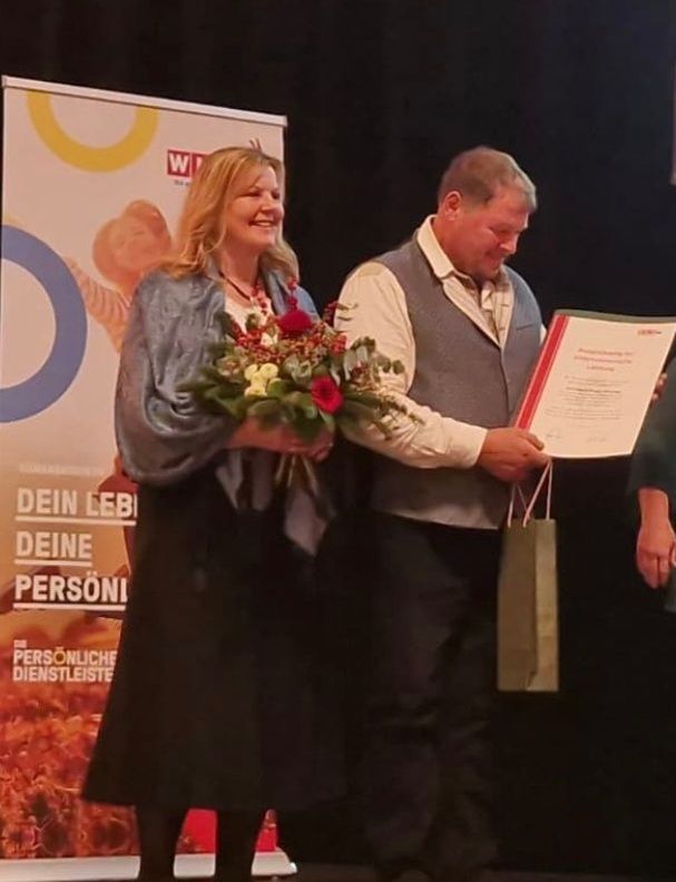 Eine Frau mit Blumenstrauß steht neben einem Mann mit Papiertüte und Urkunde auf der Bühne.