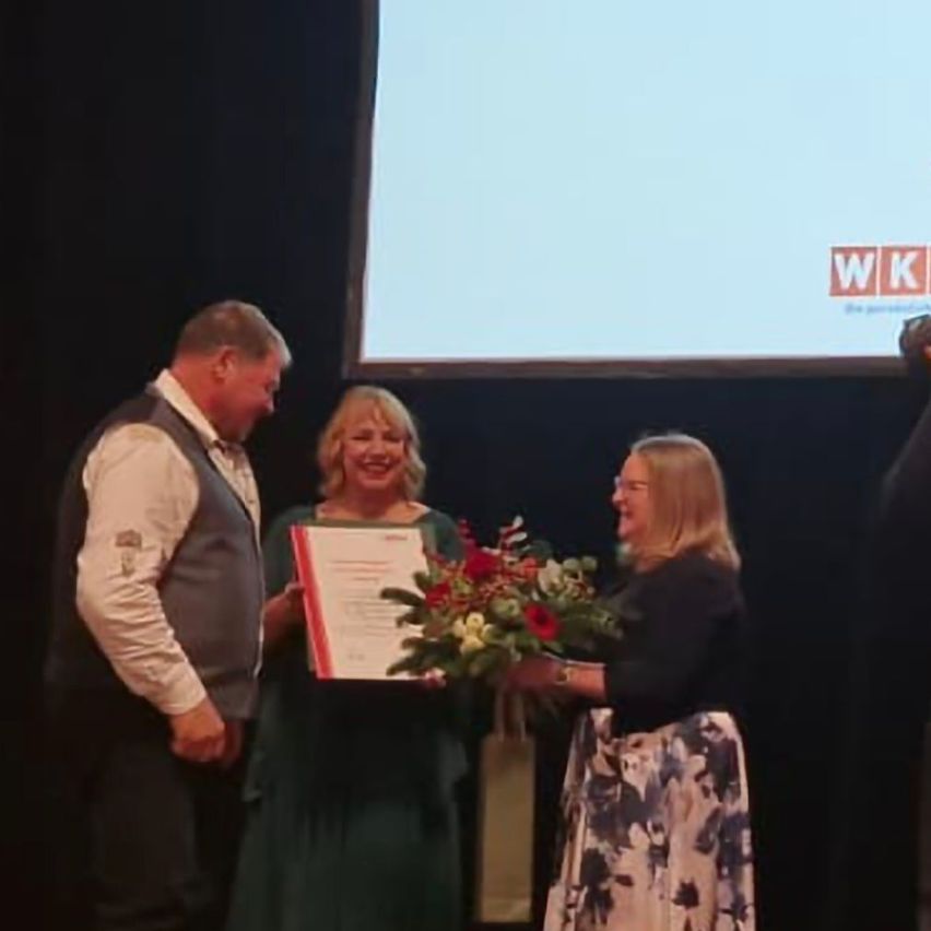 Eine Frau erhält auf der Bühne einen Preis vor einer großen Leinwand, wobei eine andere Frau Blumen überreicht.