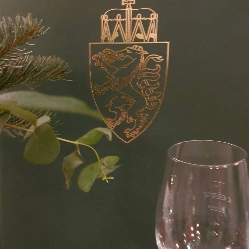 Ein goldener Schild mit Löwendesign und einem Kreuz ist auf einem dunkelgrünen Hintergrund ausgestellt. Daneben befindet sich ein kristallklares Weinglas und etwas Grün.