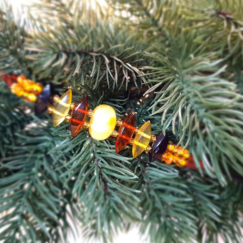 Ein farbenfroher Armreif mit gelben, orangen und dunkelblauen Perlen liegt auf einem Tannenzweig. Die Perlen sind poliert und reflektieren Licht.