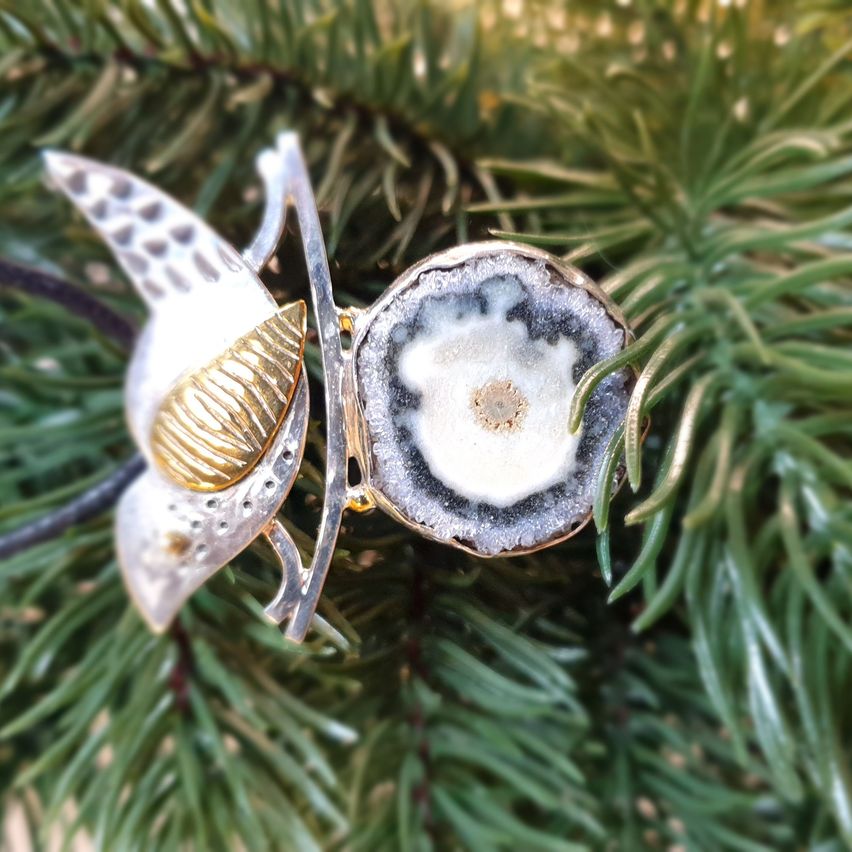 Ein silberner und goldener Schmetterlingsanhänger mit einem Achat-Zentralstein sitzt auf grünen Blättern.