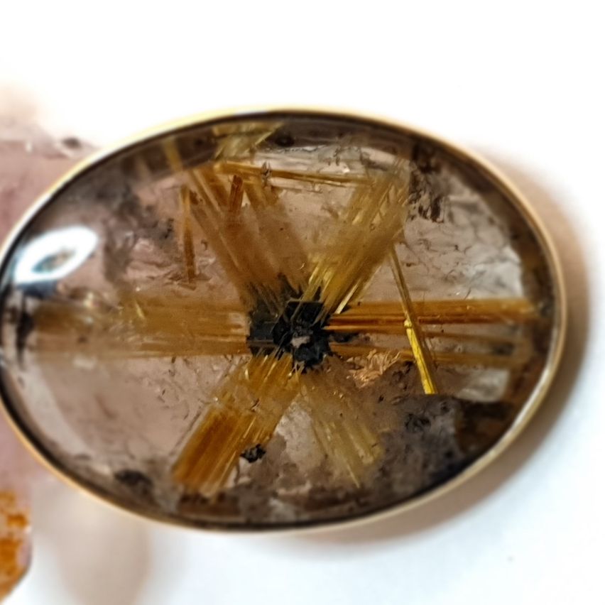 Ein golden umrandeter Kristallanhänger mit einem strahlenförmigen Muster aus goldenen Fasern, eingefasst in klarem Quarz, auf weißem Hintergrund.