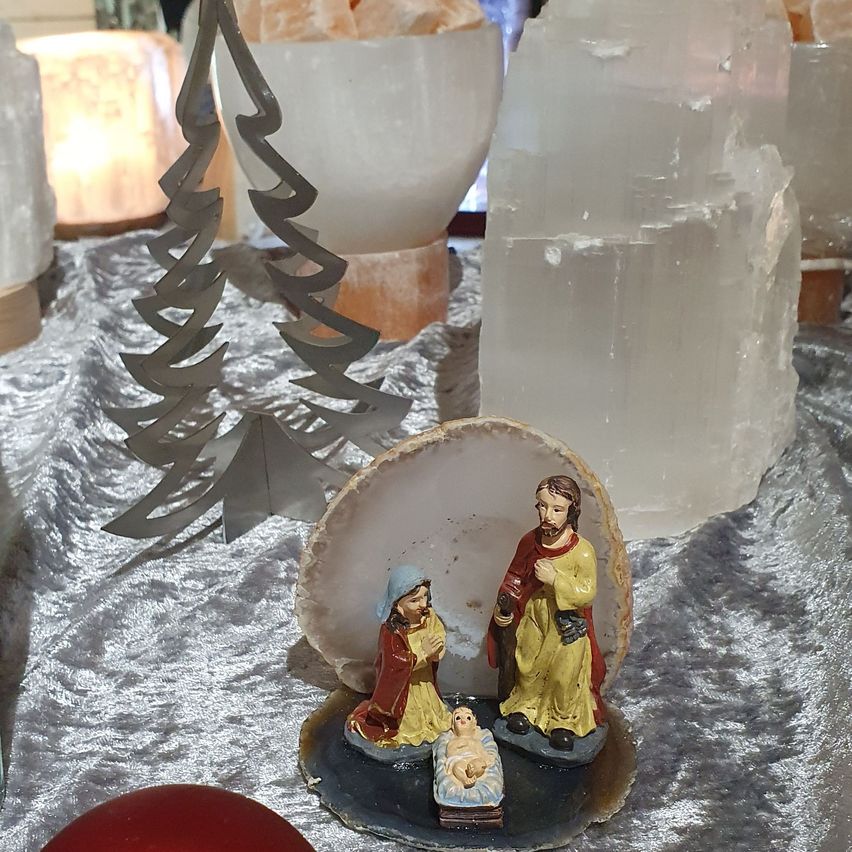 Eine kleine Krippe steht auf einem Tisch, umgeben von verschiedenen Dekorationsartikeln, darunter Kristalle und ein Miniatur-Weihnachtsbaum.