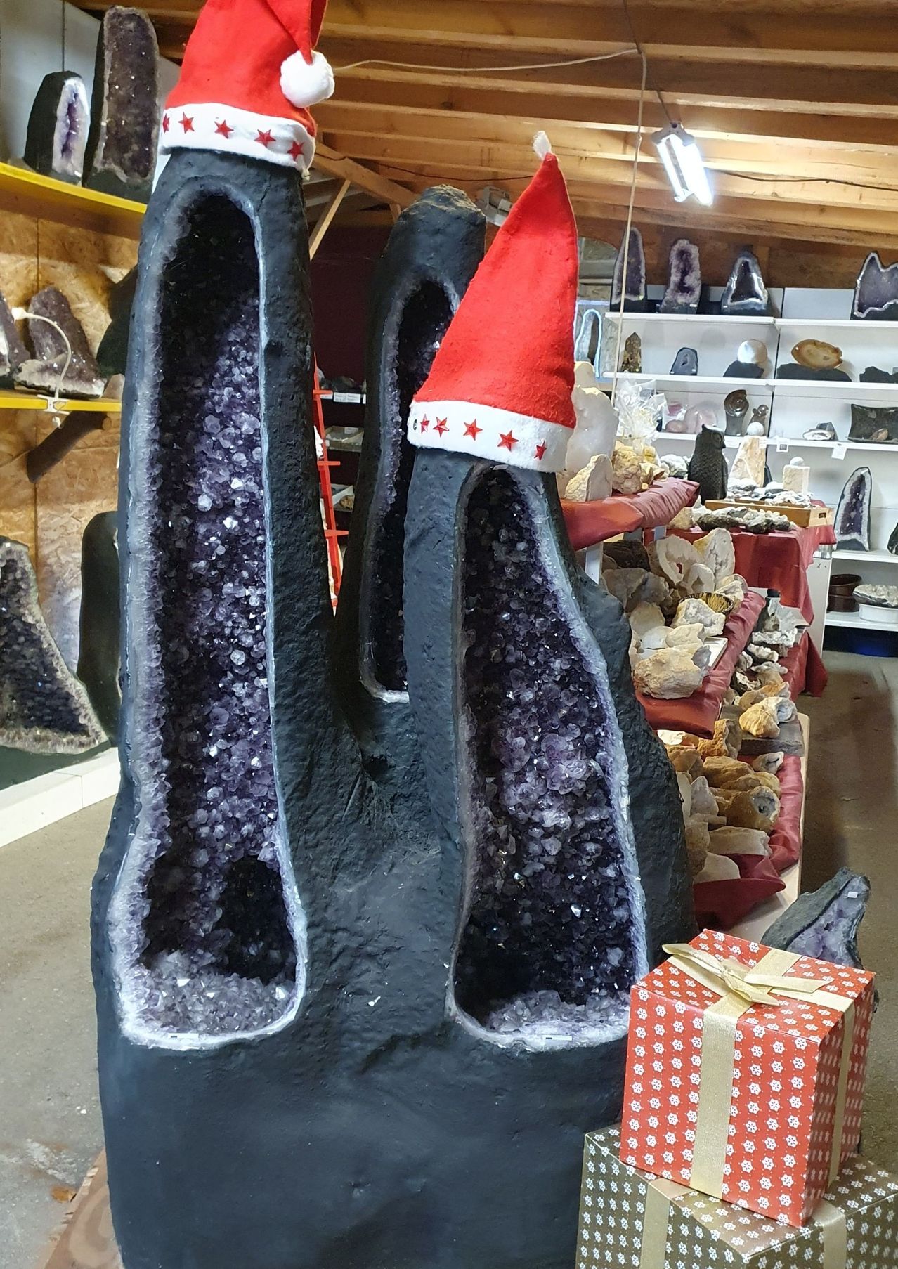 Eine Geoden-Skulptur mit Amethyst-Kristallen steht in einem Laden mit einem Weihnachtsmannhut obenauf. Dahinter zeigen Regale verschiedene Mineralien. Ein Geschenkkarton mit Schleife steht auf dem Boden.