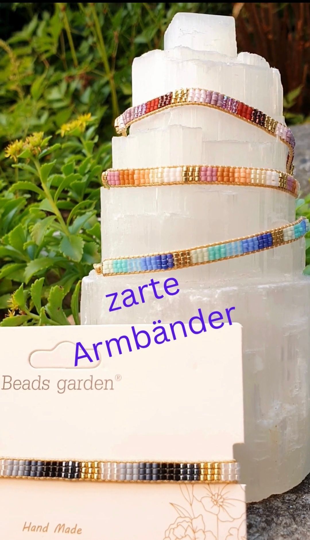 Ein Stapel bunter Perlenarmbänder mit weißem Hintergrund und dem Text 'zart Armbänder'. Die Armbänder sind vor einer Pflanze mit gelben Blüten ausgestellt.