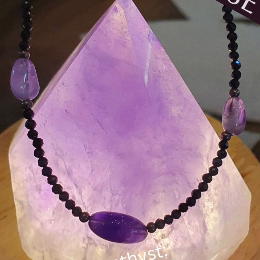 Eine Nahaufnahme eines lila Amethystkristalls mit einer Halskette aus schwarzen Perlen, die darauf ruht. Der Kristall wird auf einer Holzoberfläche ausgestellt.