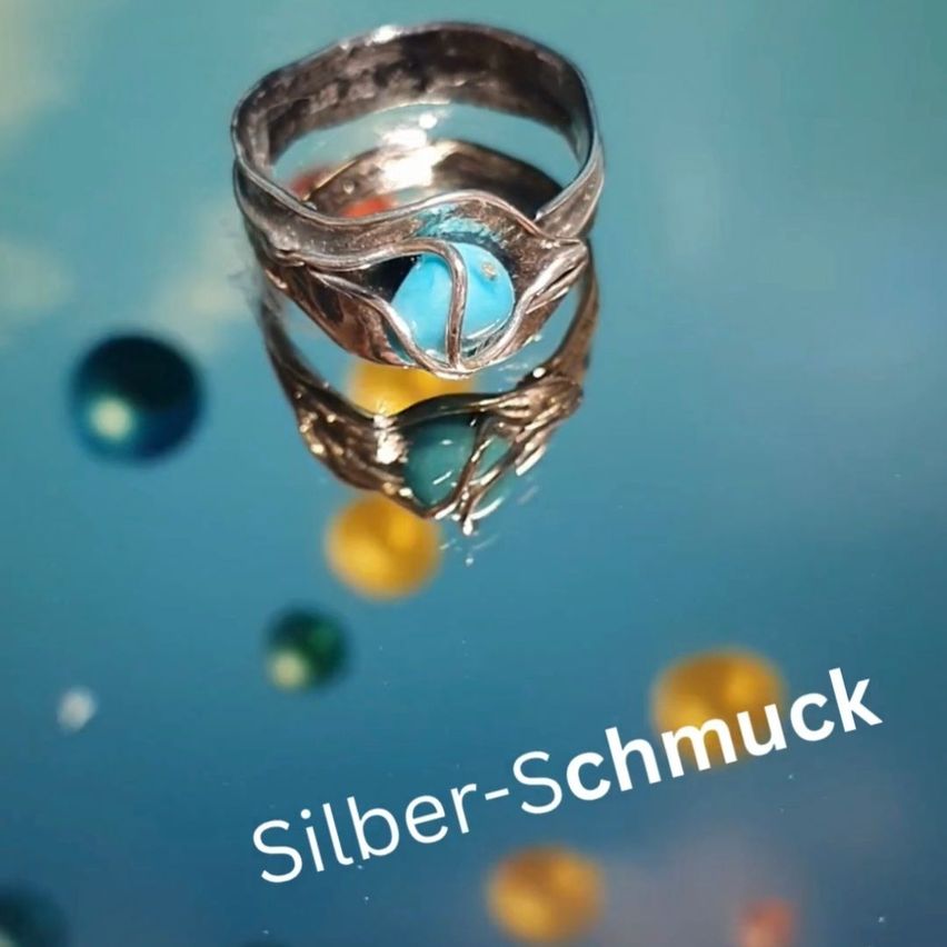 Ein silberner Ring mit türkisfarbenen Steinen schwebt über einem blauen Hintergrund mit verstreuten bunten Kugeln. Der Ring hat ein einzigartiges Design mit einem welligen Muster.