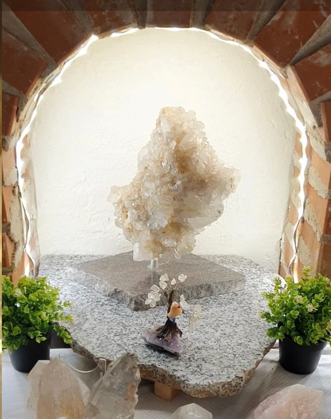 Eine große Kristallformation mit einer kleinen Figur und zwei Topfpflanzen auf einem Granitsockel, beleuchtet von einer Lichtquelle.