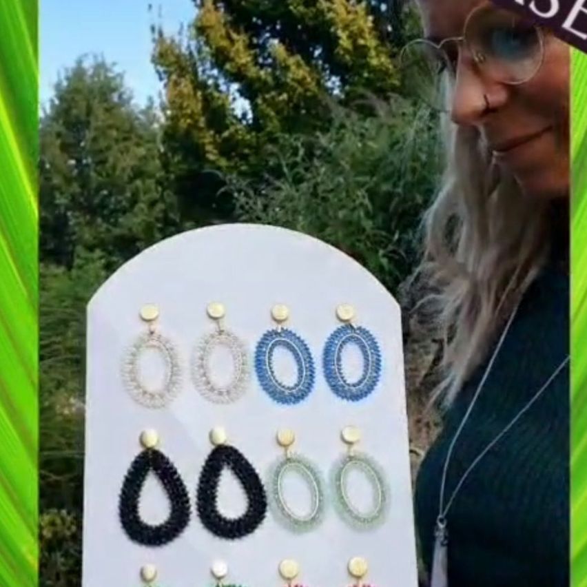 Eine Frau mit Brille hält ein weißes Brett, auf dem verschiedene gehäkelte Ohrringe in verschiedenen Farben zu sehen sind.