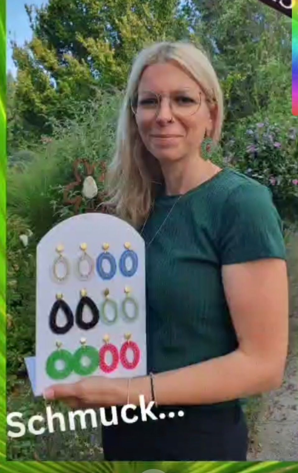 Eine Frau in einem grünen Shirt hält ein weißes Brett mit verschiedenen farbigen Ohrringen. Der Hintergrund ist ein Garten mit grünen Pflanzen und Blumen.