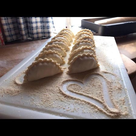 Ein Schneidebrett mit mehreren ungekochten Teigtaschen und einem Herz aus Mehl. Dahinter befindet sich ein Tablett mit einem Nudelholz.