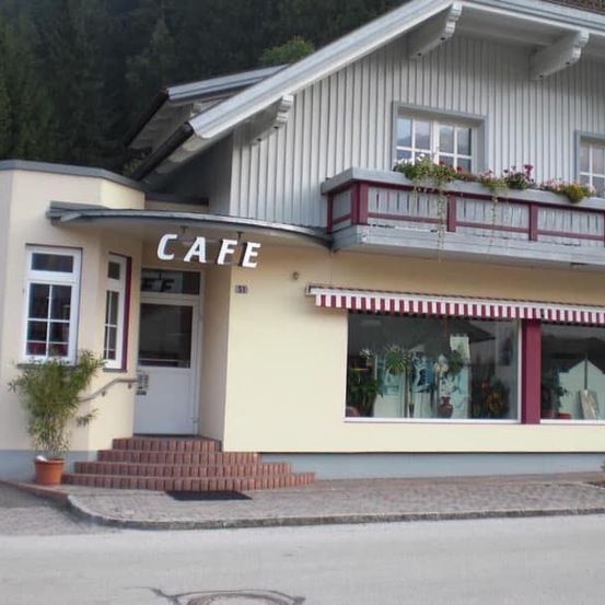 Die Vorderseite eines Cafes mit einem Balkon mit Topfpflanzen, einer weissen Tür, einem rot-weissen Vordach und einem Topf auf der linken Seite.