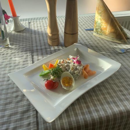 Ein Gericht mit geschnittenem Gemüse, einem halben Ei und einer Garnitur befindet sich auf einem weißen Teller auf einer gestreiften Tischdecke. Es gibt Holz-Pfefferstreuer, eine Gabel und ein Serviettentuch auf dem Tisch.