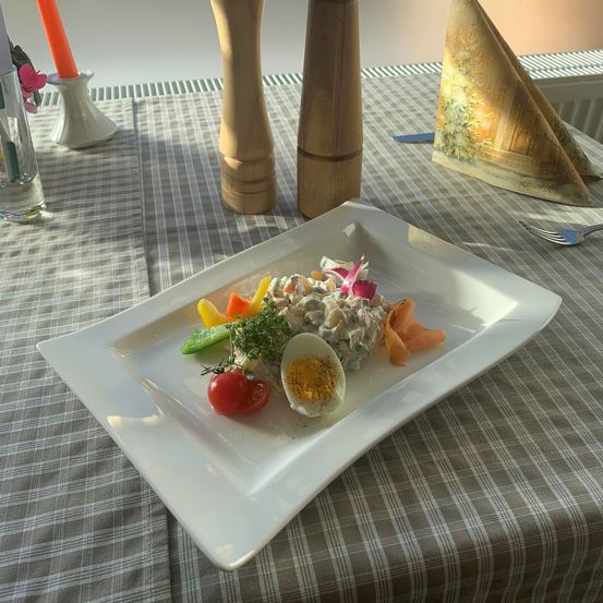 Ein Gericht mit geschnittenem Gemüse, einem halben Ei und einer Garnitur befindet sich auf einem weißen Teller auf einer gestreiften Tischdecke. Es gibt Holz-Pfefferstreuer, eine Gabel und ein Serviettentuch auf dem Tisch.