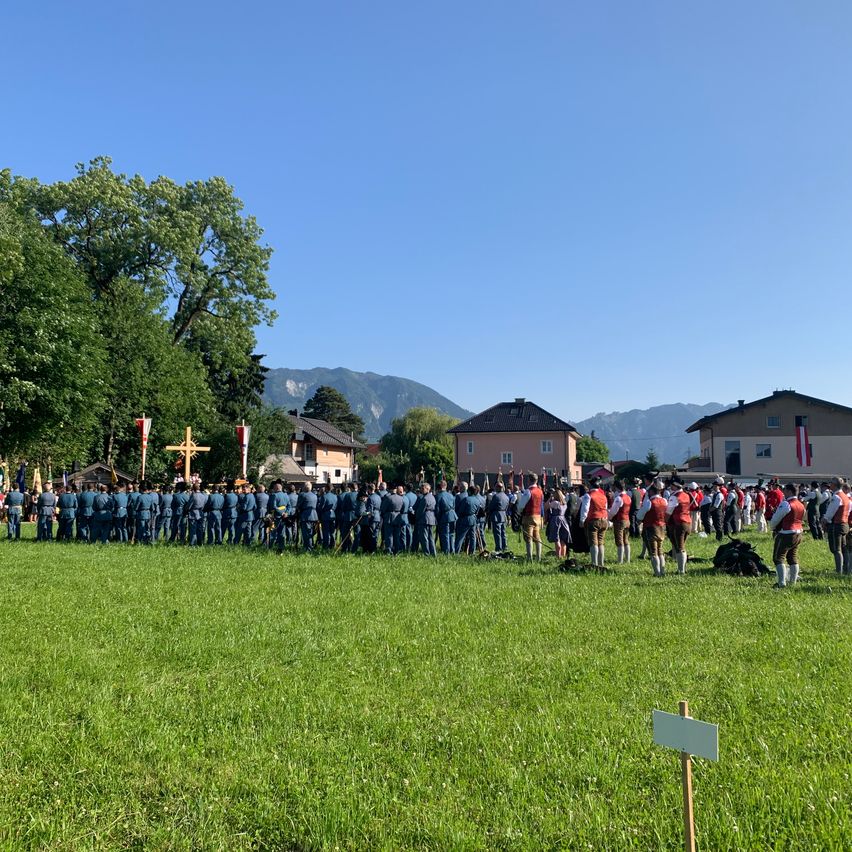 Eine große Gruppe von Menschen in Uniform steht auf einer grünen Wiese mit Bergen im Hintergrund. Einige tragen rote Westen, andere blaue. Bäume und Gebäude sind im Hintergrund.