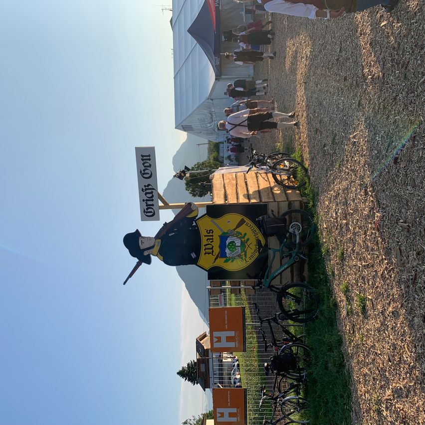 Eine Soldatenmannequin steht neben einem hölzernen Schild mit einem gelben Emblem Wache. In der Nähe sind Fahrräder auf Gras geparkt, und mehrere Leute gehen an einem Gebäude vorbei.