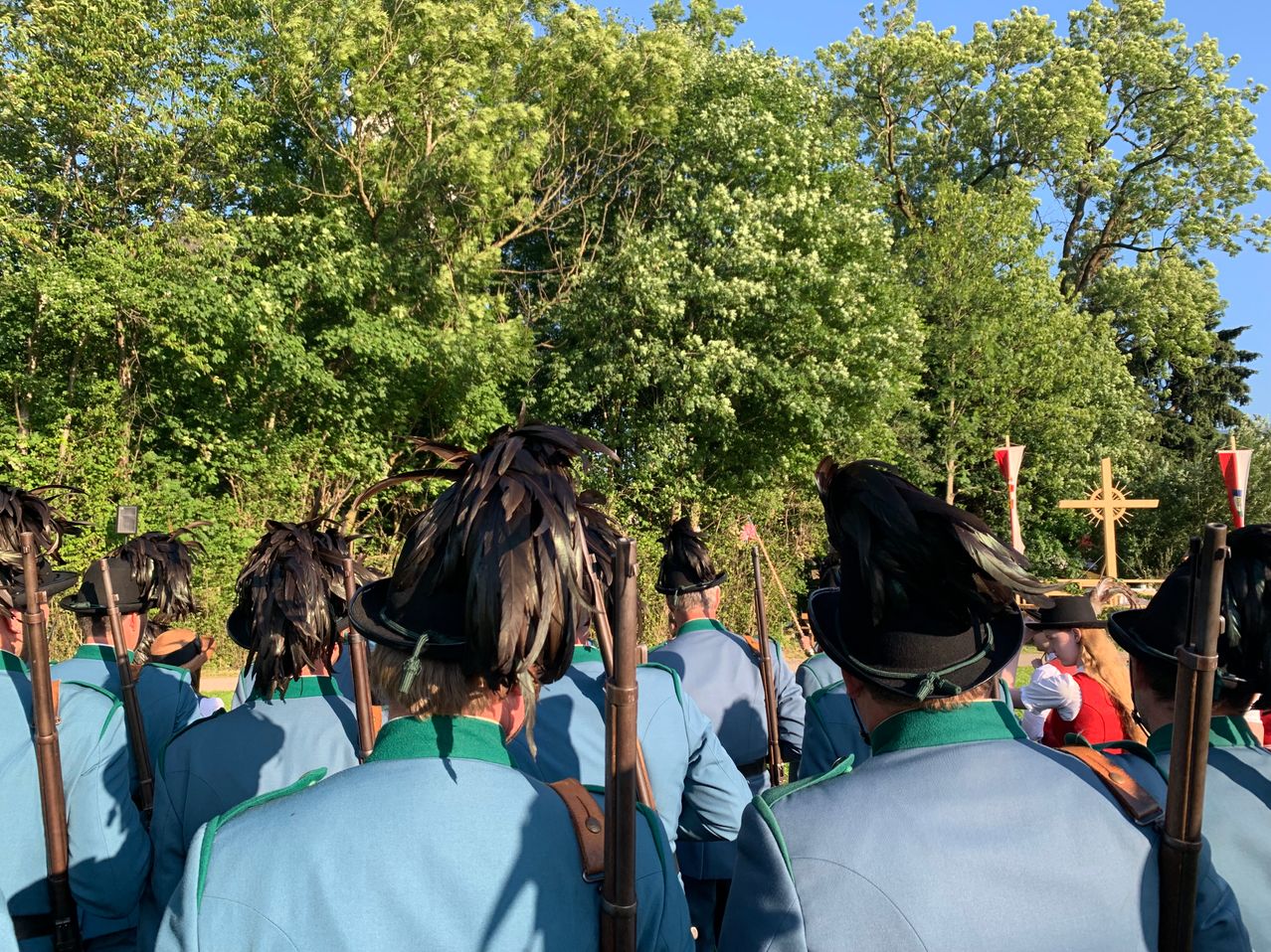 Eine Gruppe von Personen in Uniform steht in einer Reihe und hält Gewehre. Sie tragen Hüte mit Federn. Der Ort ist im Freien mit Bäumen und einer Fahne im Hintergrund.