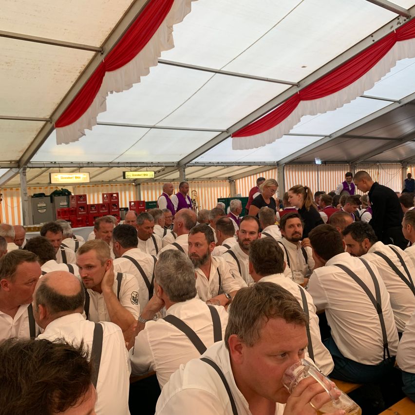 Eine Gruppe von Männern in weißen Hemden und Hosenträgern sitzt in einem Zelt mit rot-weiß gestreifter Decke. Ein Mann trinkt aus einem Bierglas.