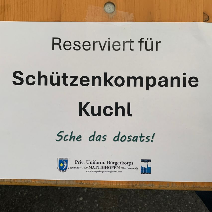 Ein weißes Schild zeigt 'Reserviert für Schützenkompanie Kuchl.' Darunter steht 'Sche das dosats!' Das Schild enthält Logos und die Website www.buergerkorps-mattighofen.com.
