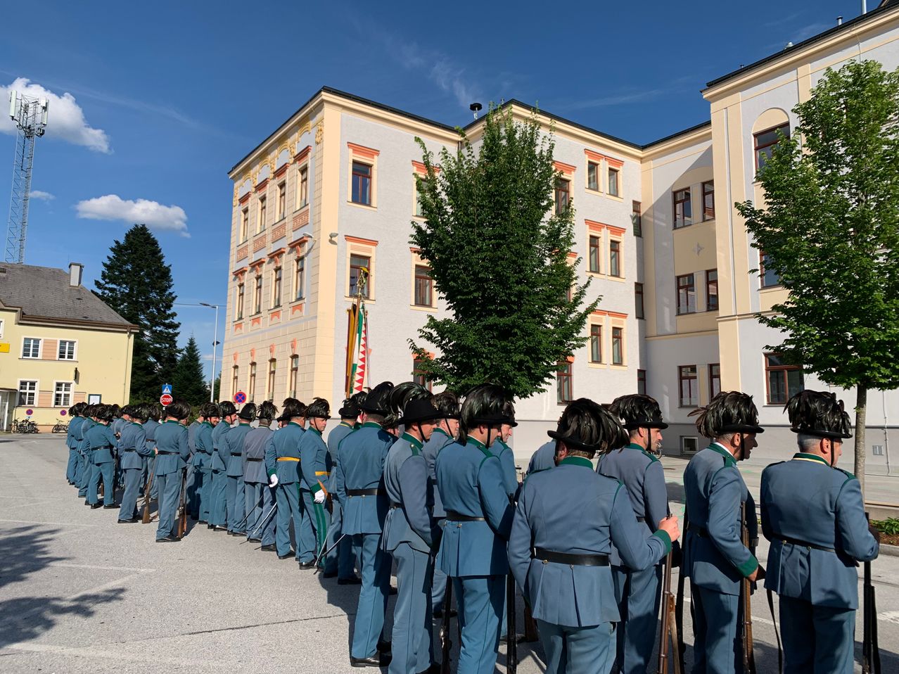 Eine Reihe uniformierter Personen steht vor einem Gebäude, wahrscheinlich für eine zeremonielle Veranstaltung. Das Gebäude weist mehrere Fenster und dekorative Elemente auf.