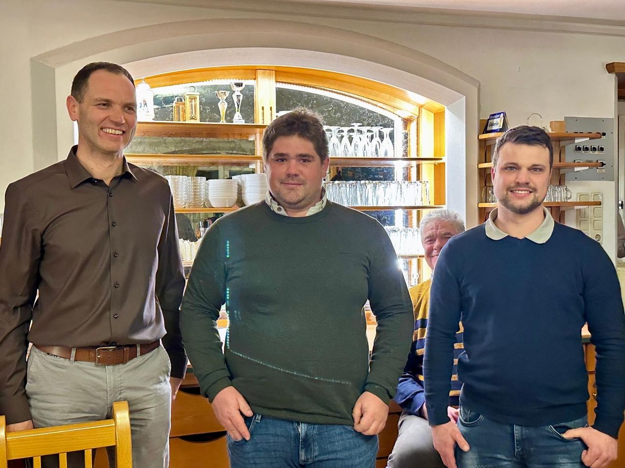 Drei Männer posieren für ein Foto in einem Raum mit einem Schrank und einem sitzenden älteren Mann. Der Mann in Grün hat seine Hände in den Taschen.