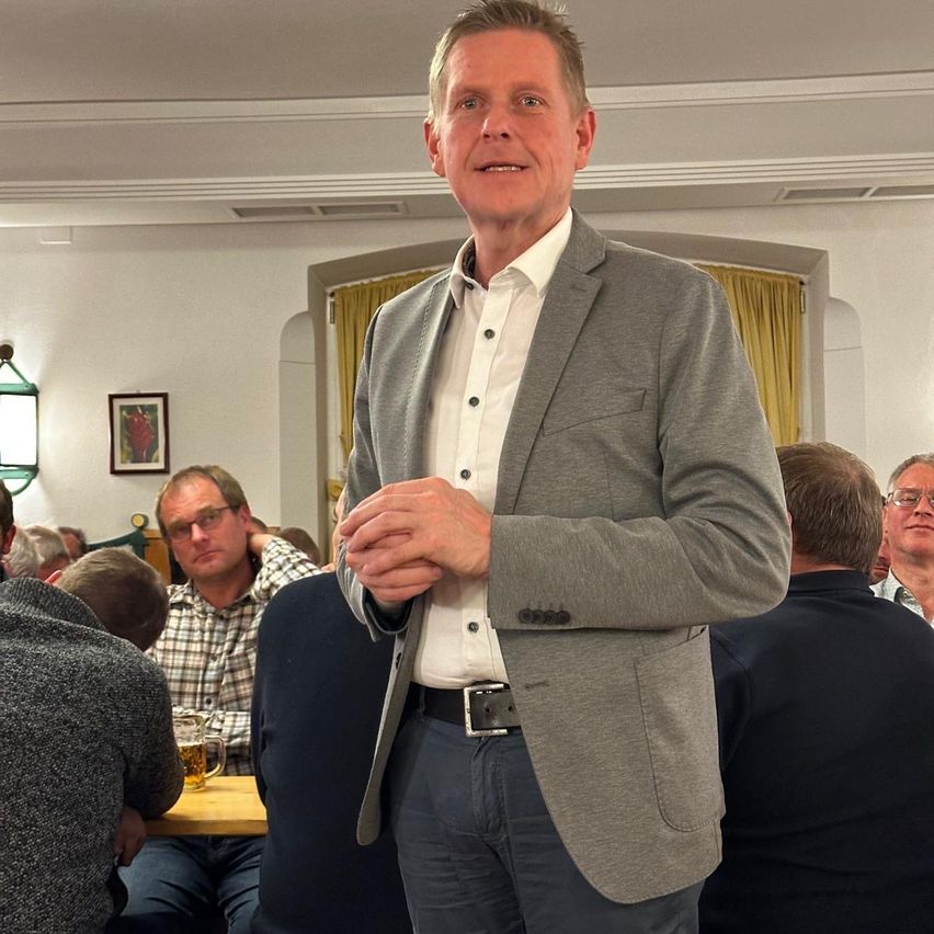 Ein Mann in einem grauen Blazer und weißem Hemd steht vor einem sitzenden Publikum und lächelt. Hinter ihm sitzt ein Mann in einem karierten Hemd mit einem Glas Bier auf einem Tisch. Ein weiterer Mann mit Brille sitzt rechts. Ein Bilderrahmen hängt an der Wand.